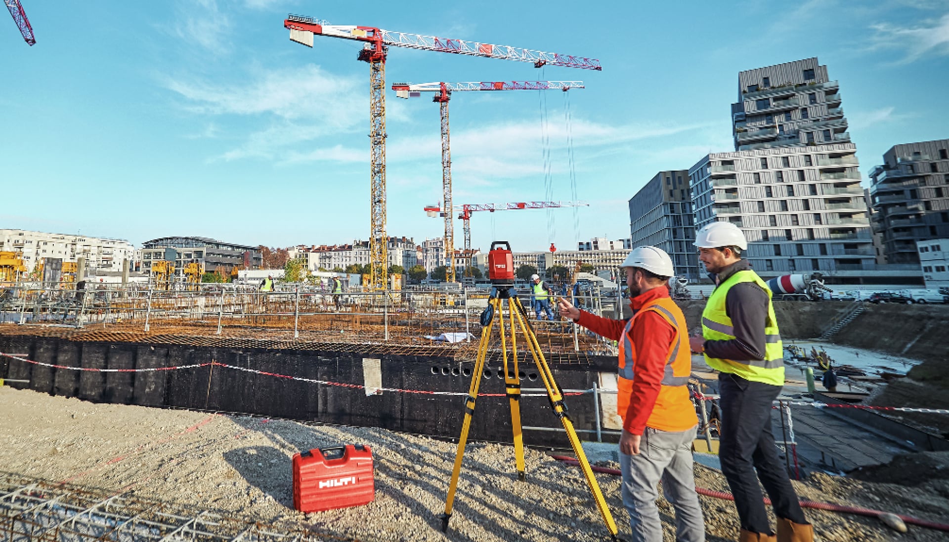 Two men stood infront of a tripod on a construction site with cranes in the background