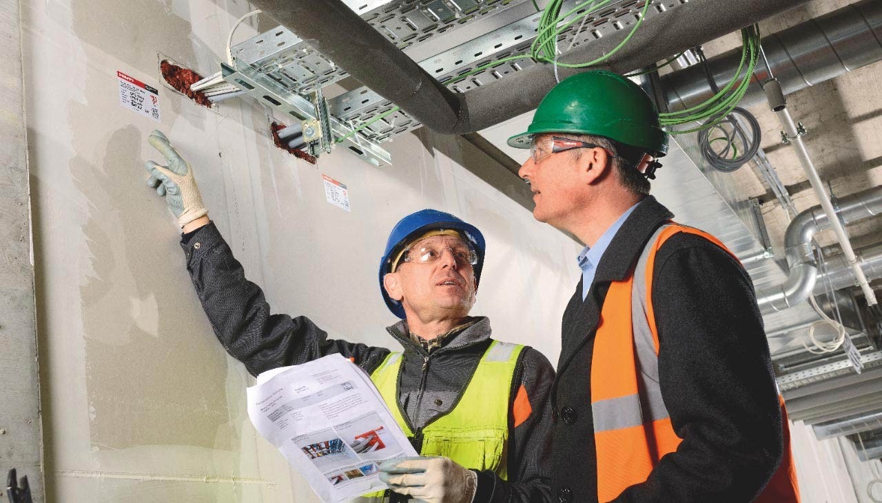 2 men in hard hats, one man holding a document and pointing towards a sticker on a wall
