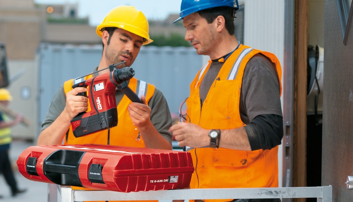 2 men in hard hats and hi vis vests with 1 holding a Hilti red drill